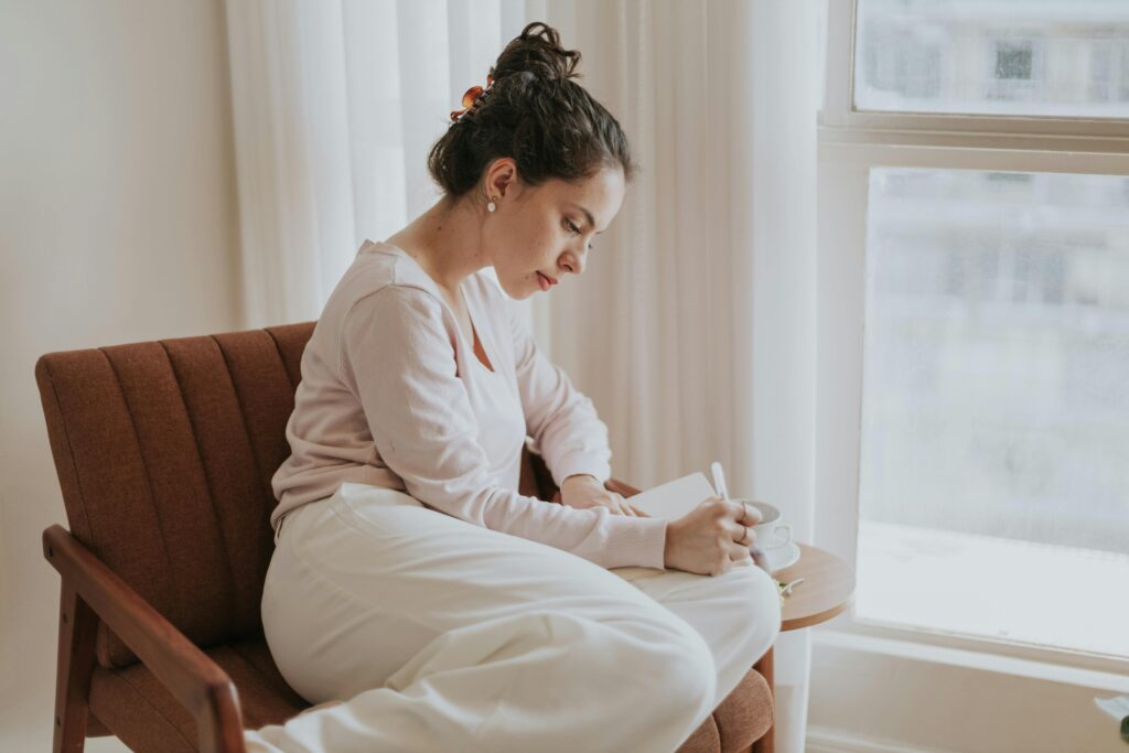 pexels-photo-33359325-33359325 A woman comfortably journaling by a window, surrounded by soft natural light, in a cozy indoor setting.