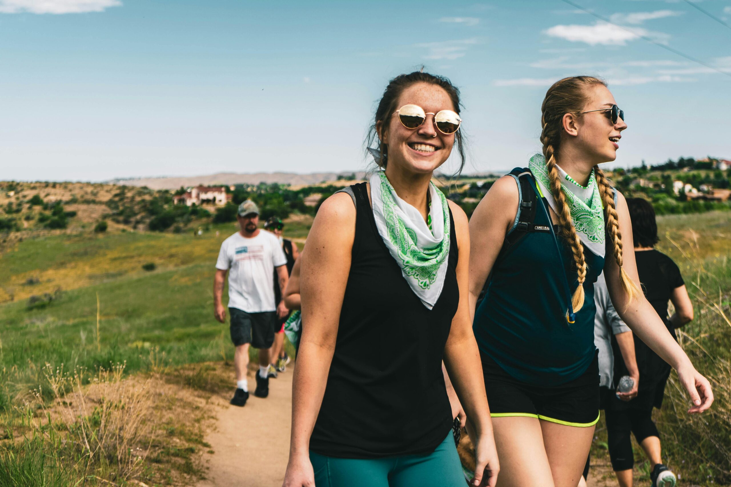 A group of adults hiking on a sunny day in a grassy landscape, wearing bandanas and sunglasses.