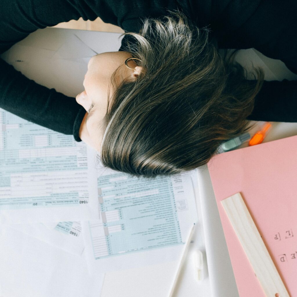 pexels-photo-6919765-6919765 Exhausted woman resting her head on a desk cluttered with papers and a ruler, showing workplace fatigue.